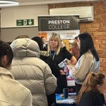 People gathered around the Preston College stand at a jobs fair, speaking with staff and browsing course information.